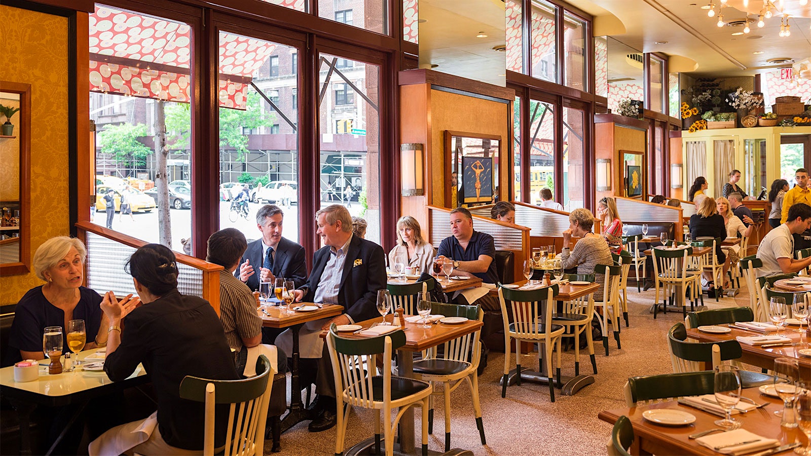 People dining in the dining room at Nice Matin, with modern-design posters on the wall and white wooden chairs with black seats, with windows looking onto the Upper West Side